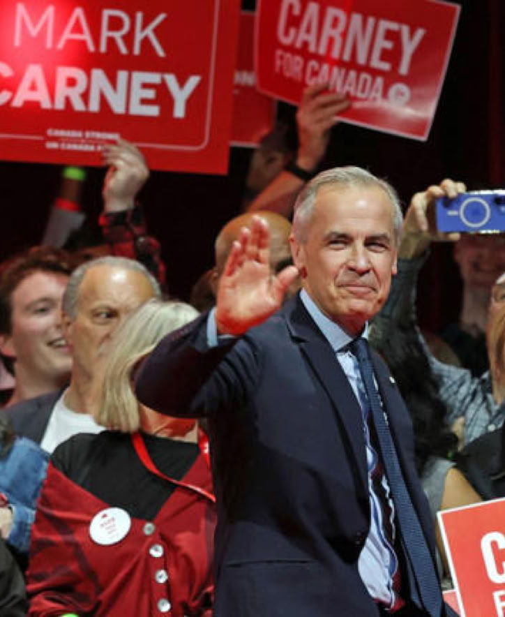 A group of people holding red signs with text, Mark Carney in the center waving, surrounded by others smiling and clapping.