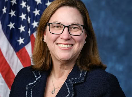 A woman with glasses and shoulder-length hair is smiling, wearing a dark blazer with white trim. An American flag is visible in the background.
