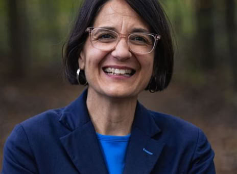 A person with short dark hair and glasses, wearing a blue shirt and blazer, smiling in an outdoor setting with blurred trees in the background.