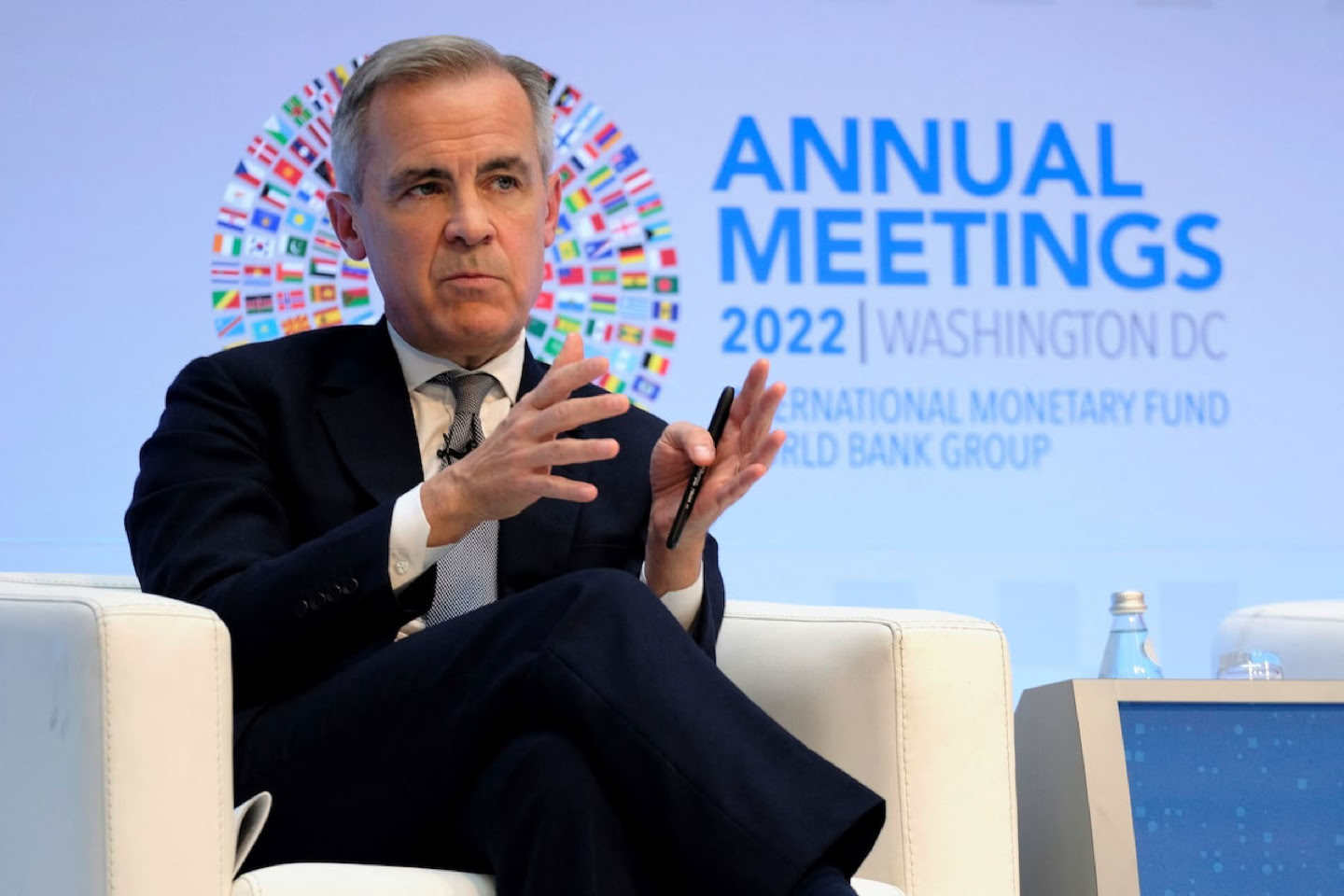 Mark Carney in a suit speaking on stage with "Annual Meetings 2022 Washington DC" text and logos in the background.